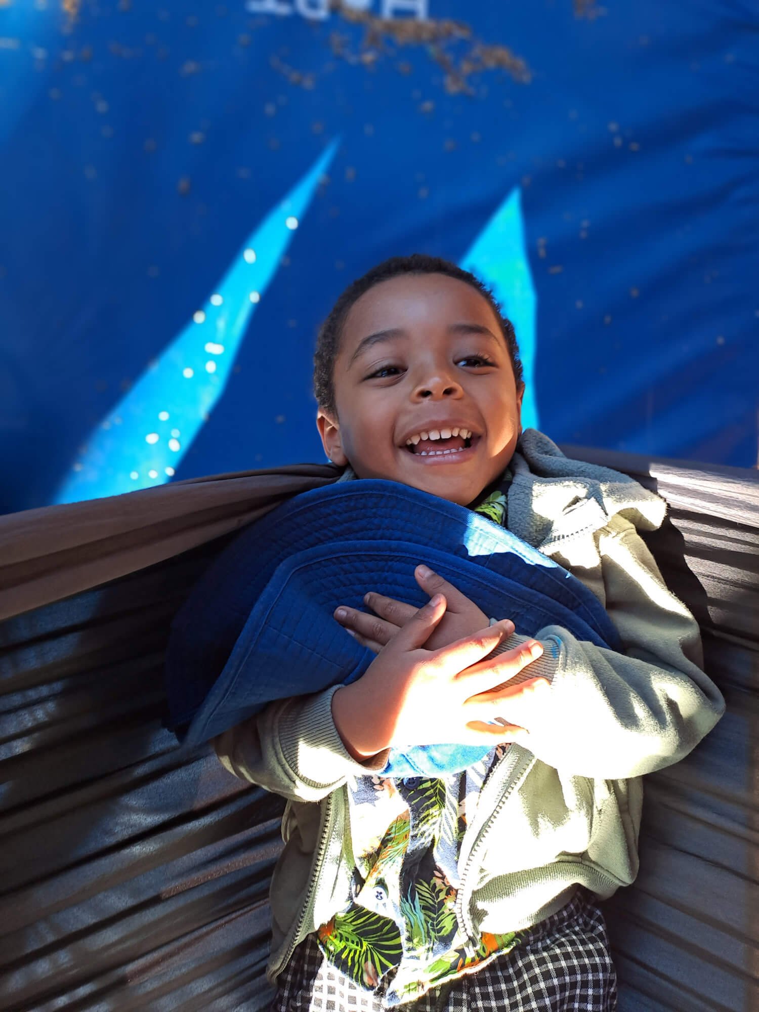 Smiling young boy lying on a hammock with a blue background featuring a light pattern. His hand rests on his chest, and he is wearing a beige jacket and patterned shorts.