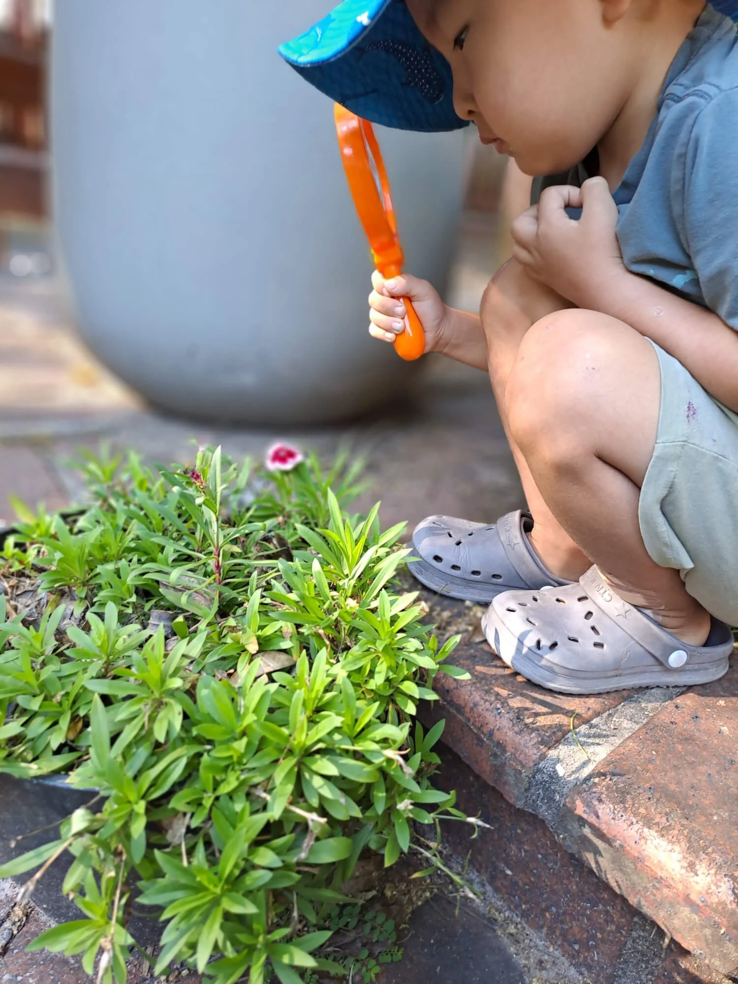 A young child crouching on a brick sidewalk, holding an orange bubble wand, blowing bubbles near a small bush with pink and white flowers. The child is wearing grey slip-on shoes, khaki shorts, and a grey t-shirt, with a blue hat.