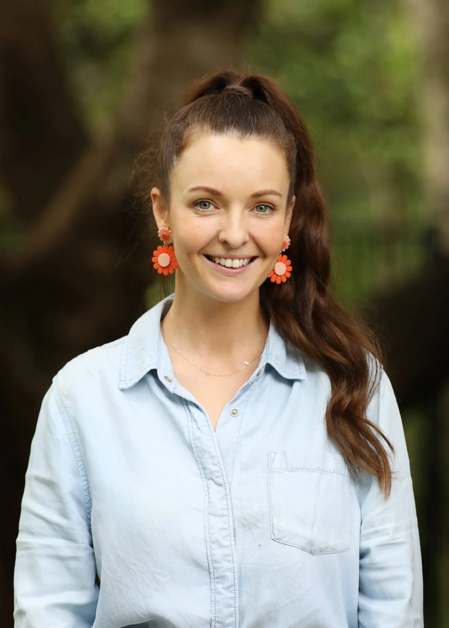 Smiling woman outdoors with brown hair in a high ponytail, wearing large orange flower earrings and a light blue button-up shirt.