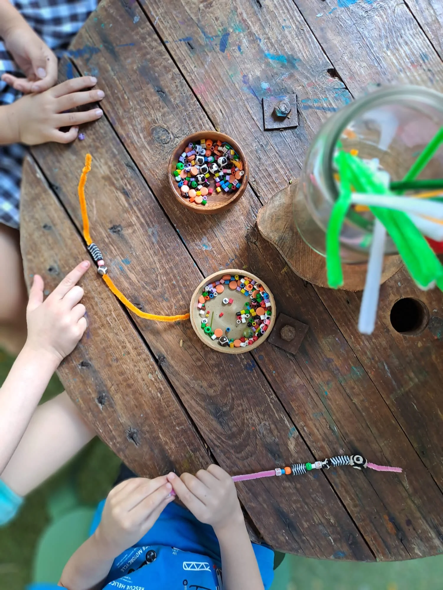 Two children at Ballymore Kindy, Herston, are making jewellery with colourful beads and pipe cleaners on a rustic wooden table, with a jar of straws nearby.