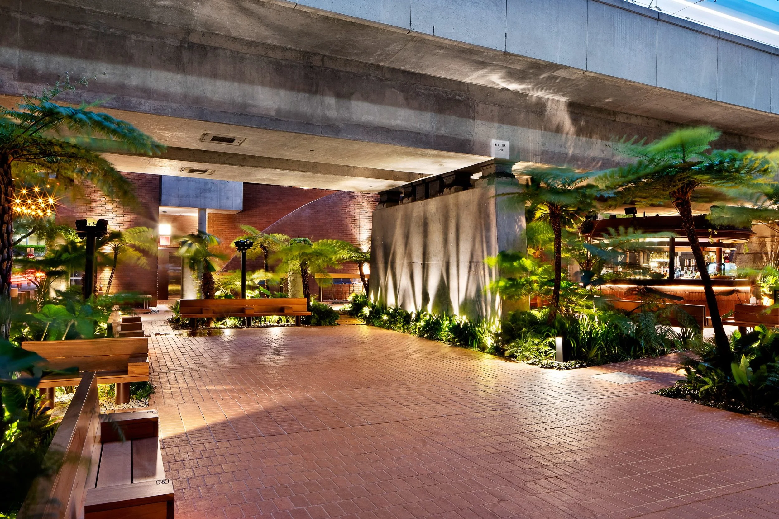 An urban outdoor scene featuring a brick-paved area with wooden benches, lush green trees, and plants illuminated by warm lighting under a large concrete overpass.