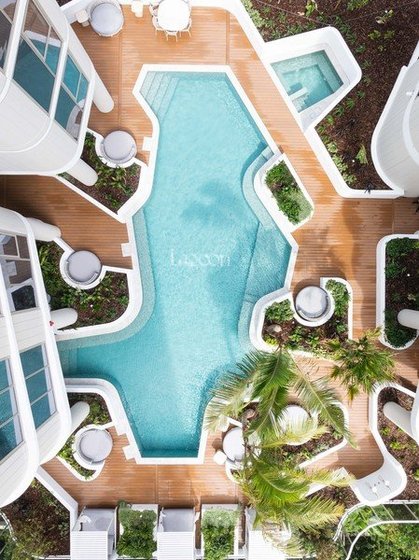 Bird's eye view of a hotel swimming pool area with wooden deck, surrounded by white lounge chairs, umbrellas, and tropical plants.