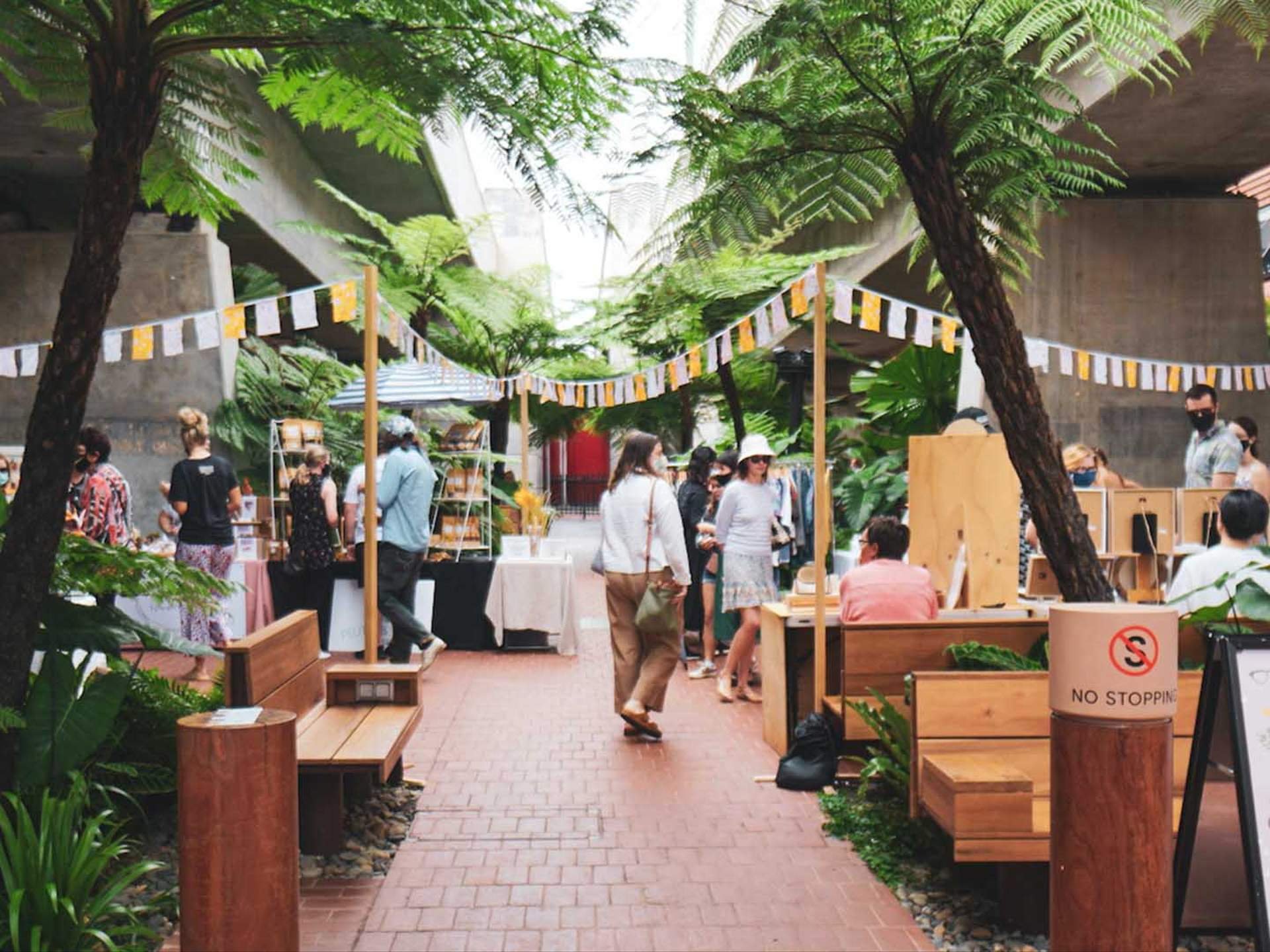 People shopping and browsing at an outdoor market under large green trees and colorful banners, with wooden stalls and greenery.