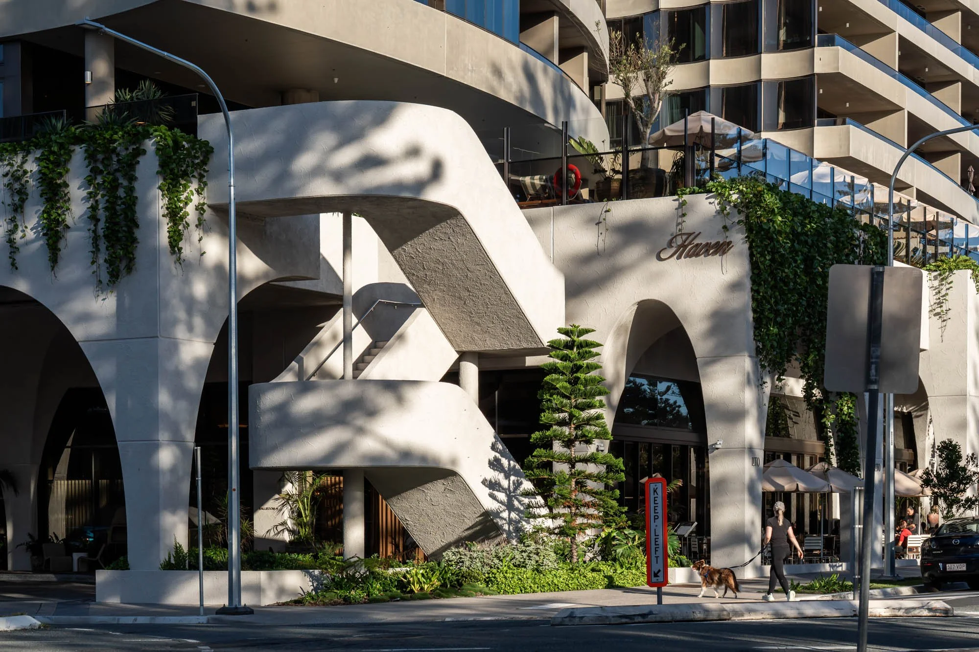 Modern multi-story building with curved balconies, greenery, and outdoor seating areas. Pedestrians walking a dog on the street in front. Sign indicating no parking on the curb.
