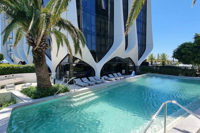 Modern hotel poolside with a palm tree, lounge chairs, and a high-rise building with reflective windows in the background.