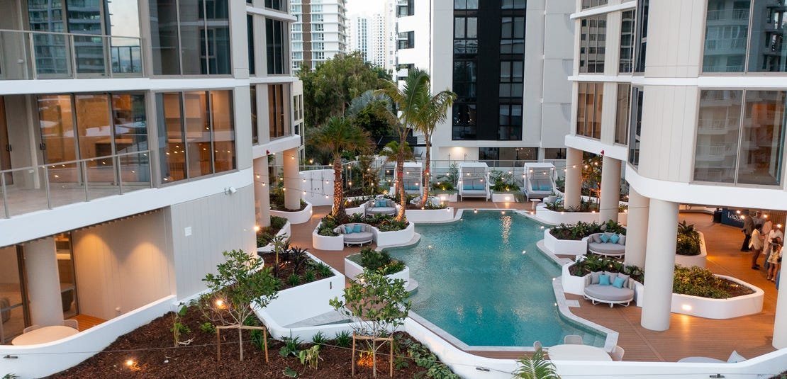 Apartment complex courtyard with a pool surrounded by lounge chairs, palm trees, and modern high-rise buildings.