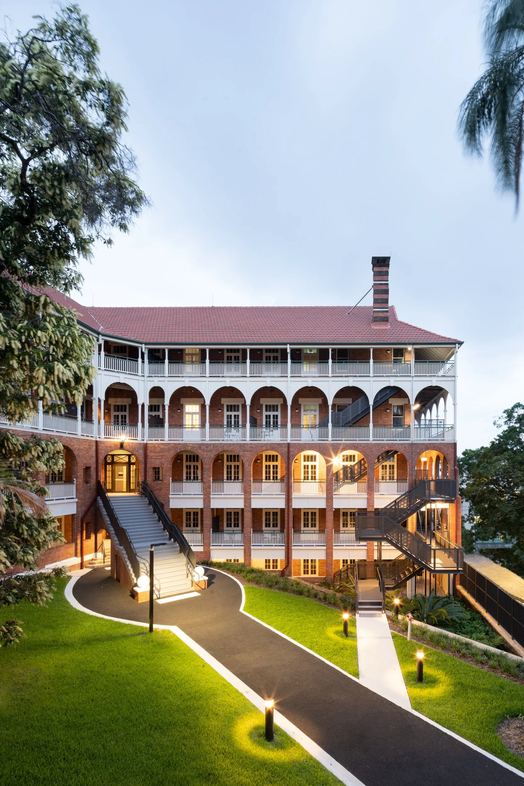 A multi-story brick building with white railings and arched doorways, illuminated with exterior lighting, surrounded by a landscaped yard with green grass, walking paths, and outdoor stairs.