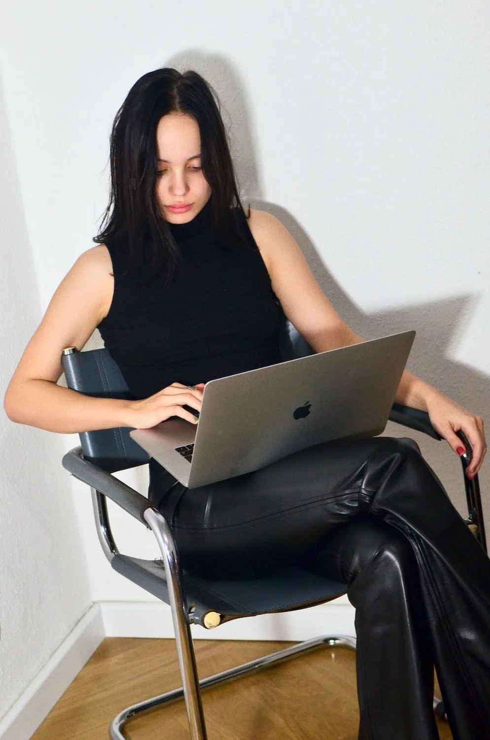 A young woman with black hair, wearing a black sleeveless top and black leather pants, sitting on a black chair with metal legs. She is working on a silver MacBook laptop, with her left hand on the keyboard and her right hand resting on the armrest, in a room with white walls and wooden flooring.