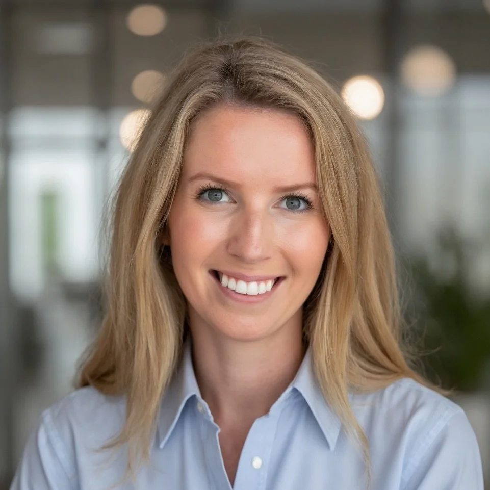 A young woman with blonde hair and blue eyes, smiling, wearing a light blue button-up shirt, in a modern indoor setting with blurred background lights.