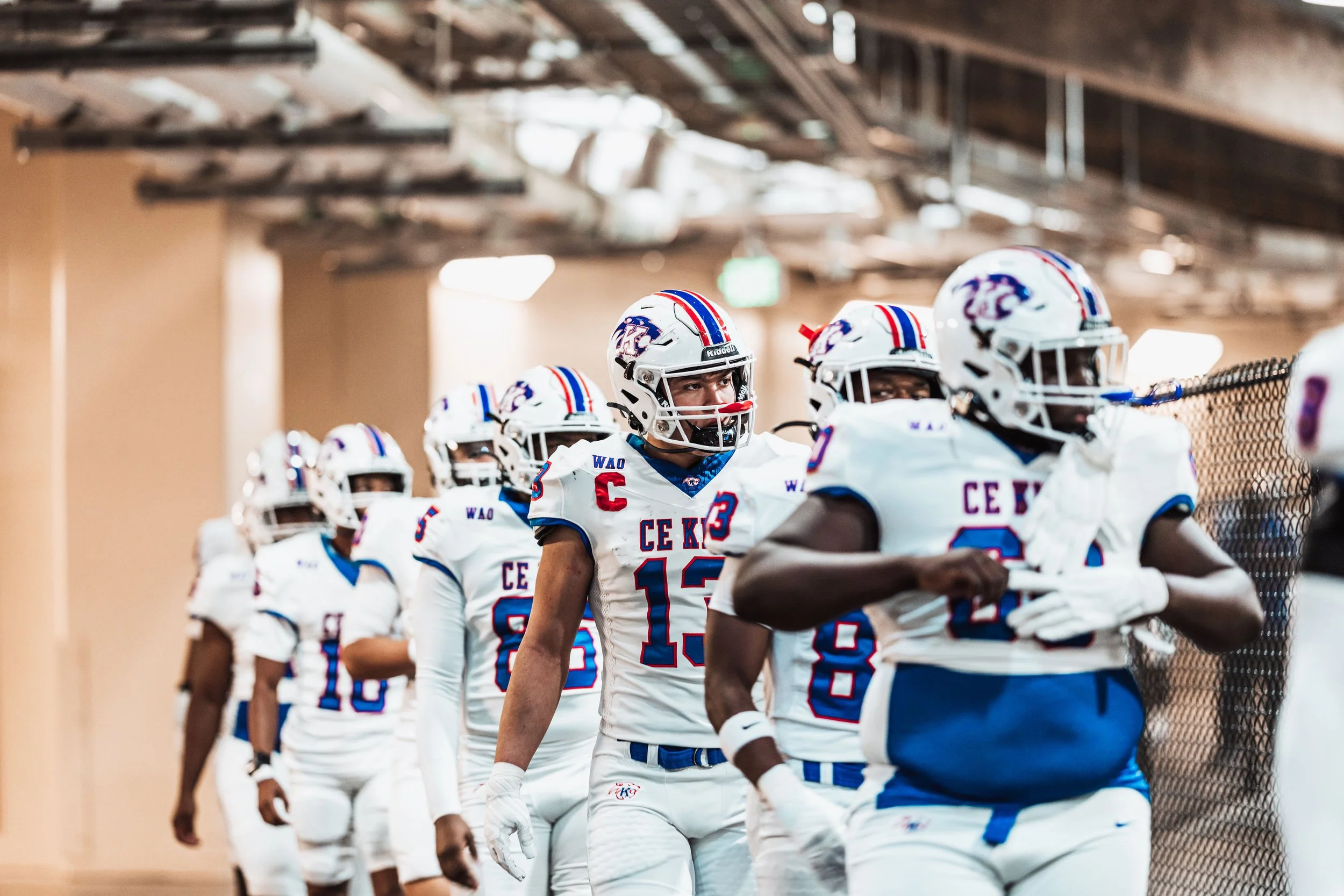 A group of American football players in white uniforms with blue and red accents walking in a line in a tunnel.