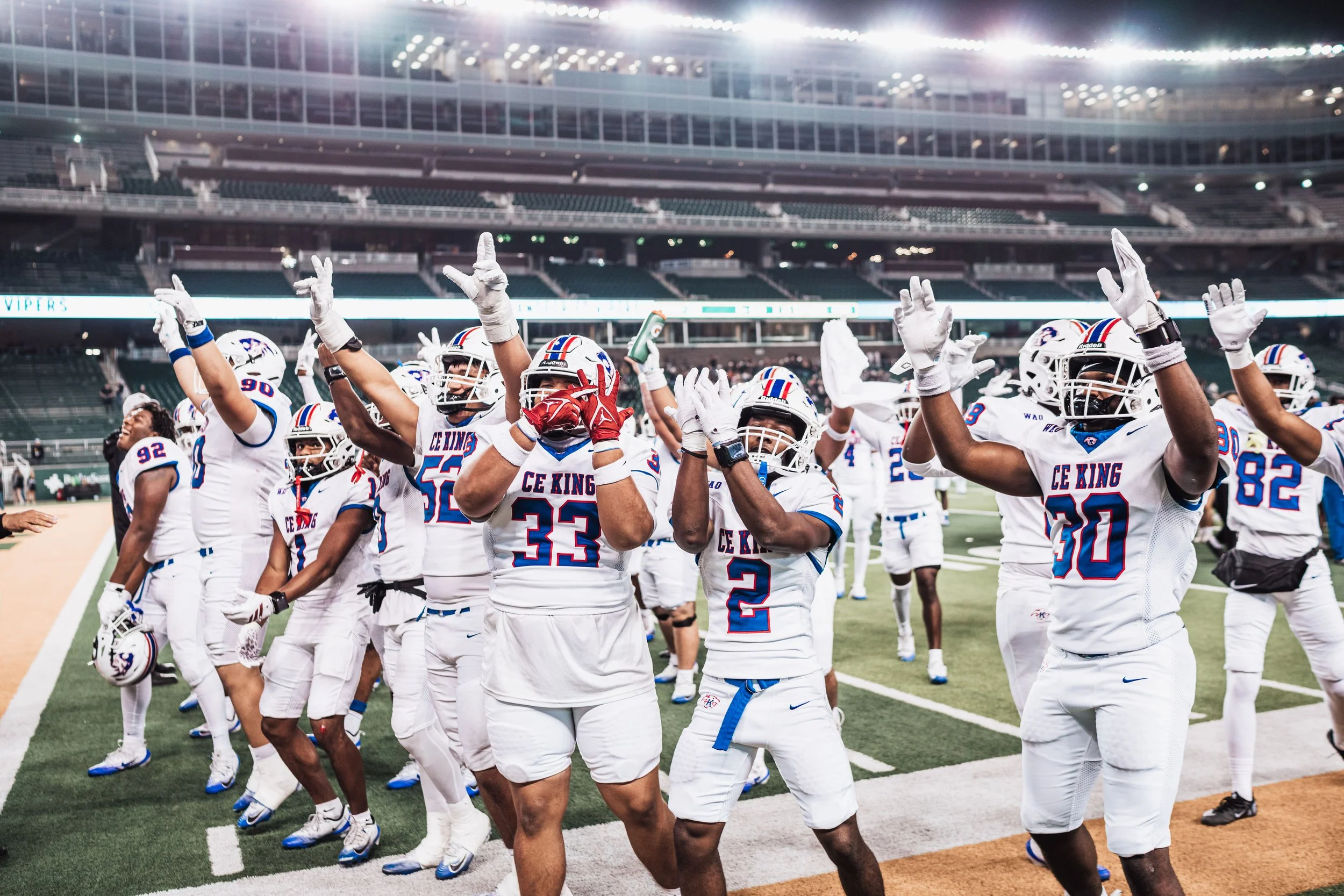 A group of football players in white uniforms with red and blue accents celebrating on the field inside a stadium with bright lights, some raising their hands and others clapping.