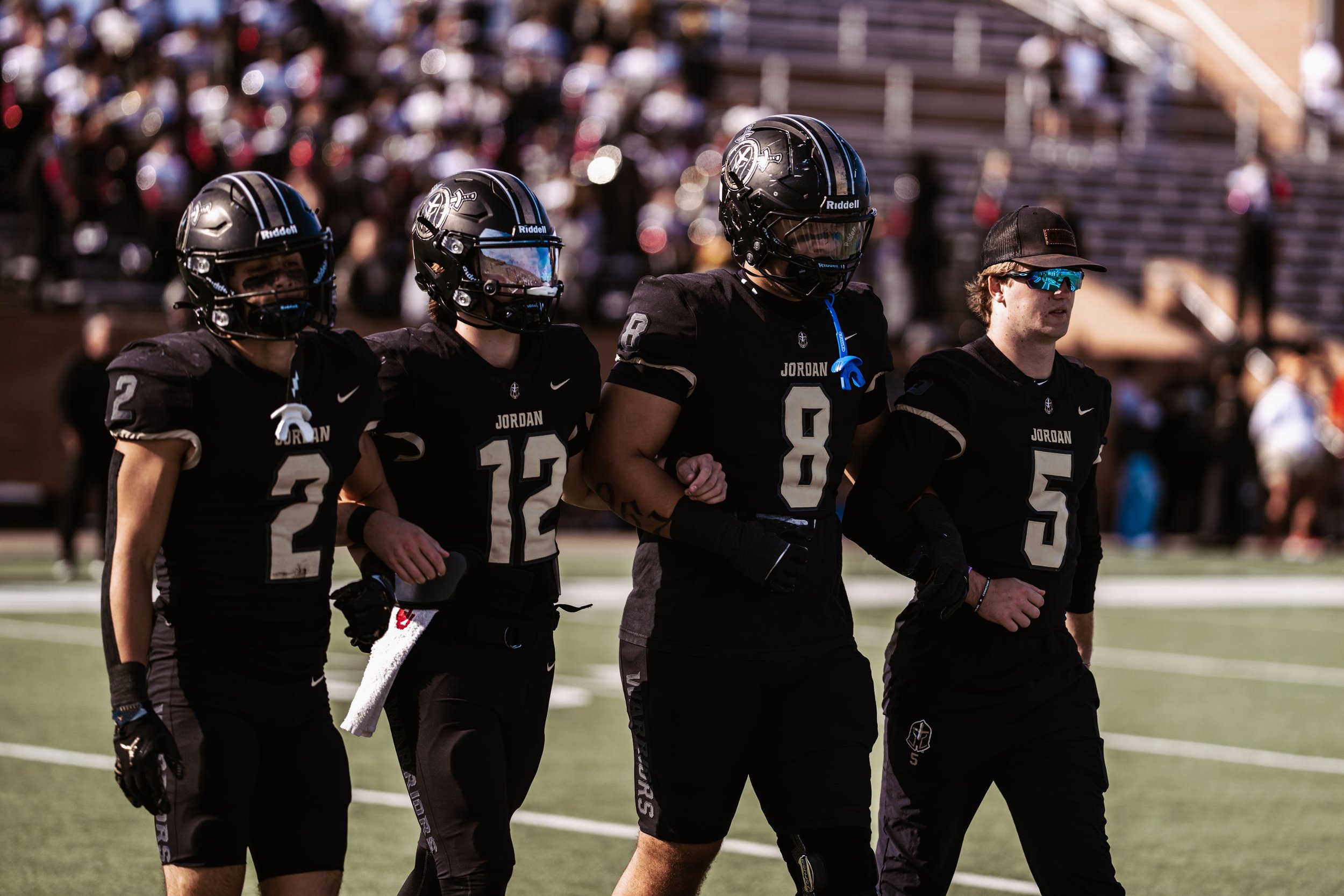Four football players in black uniforms and helmets walking on the field with a blurred crowd in the background.