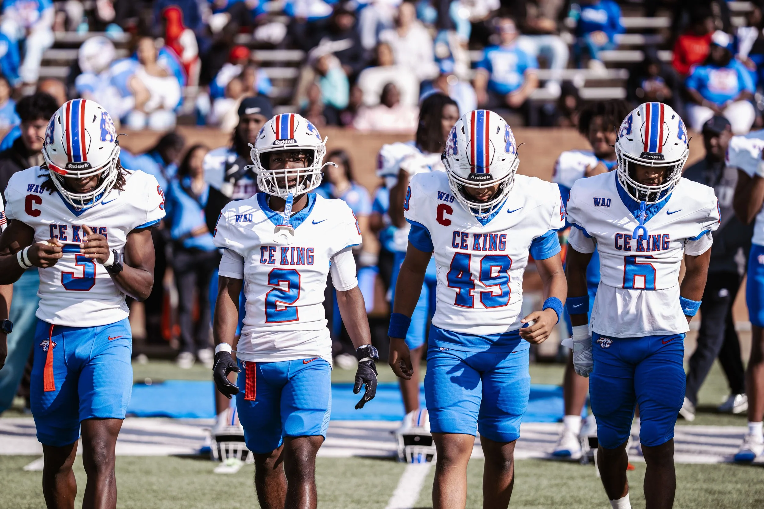 Four young football players wearing white and blue uniforms with helmets on, walking on a football field during a game. Fans sitting in the background.