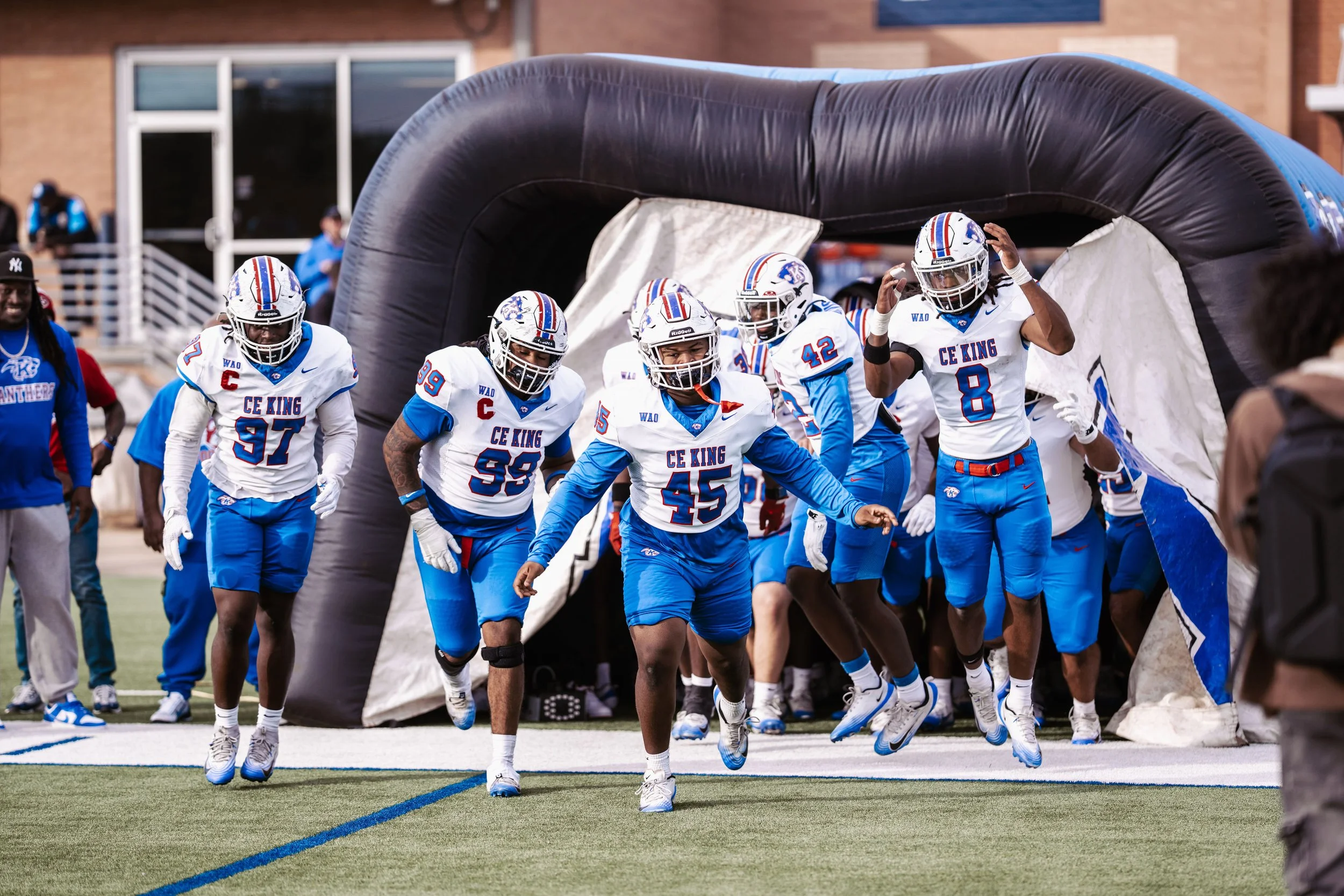A group of football players in blue and white uniforms, with some wearing helmets, running out of a dark inflatable tunnel onto the field during a game.