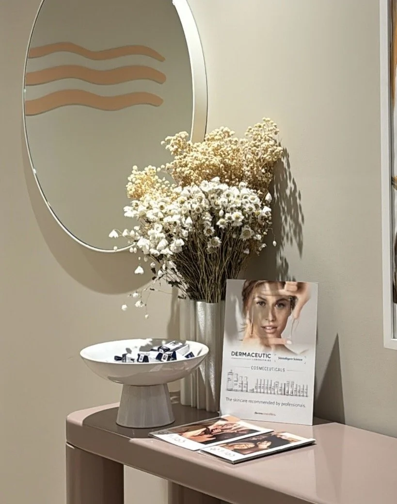 A vanity table setup with a tall vase of dried white flowers, a bowl filled with small bottles or containers, and skincare informational brochures. There's a round mirror on the wall and a poster behind the flowers featuring a woman's face and skincare products.
