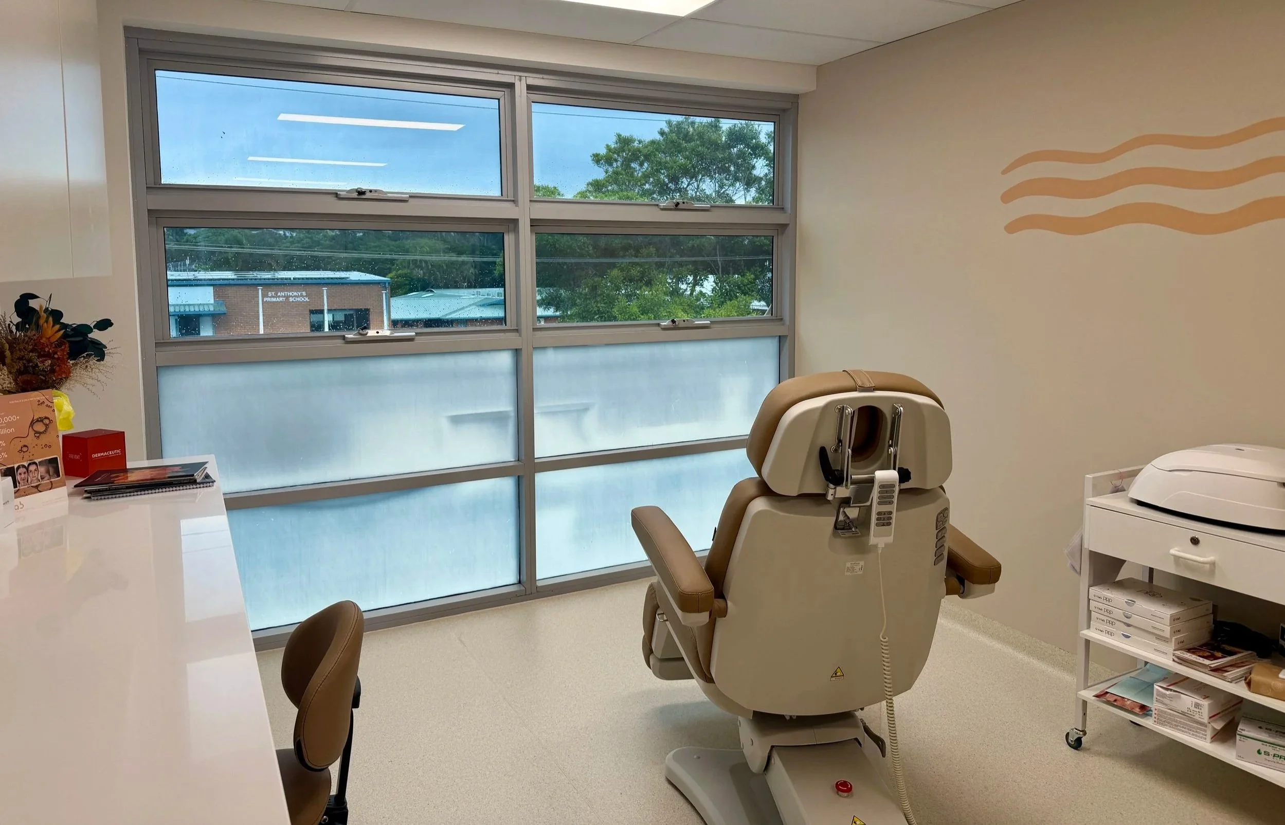 A medical treatment room with a beige examination chair, a white medical device, a small white table with supplies, and a large window showing an outside building labeled 'St. Anthony's Primary School' with trees and a cloudy sky.
