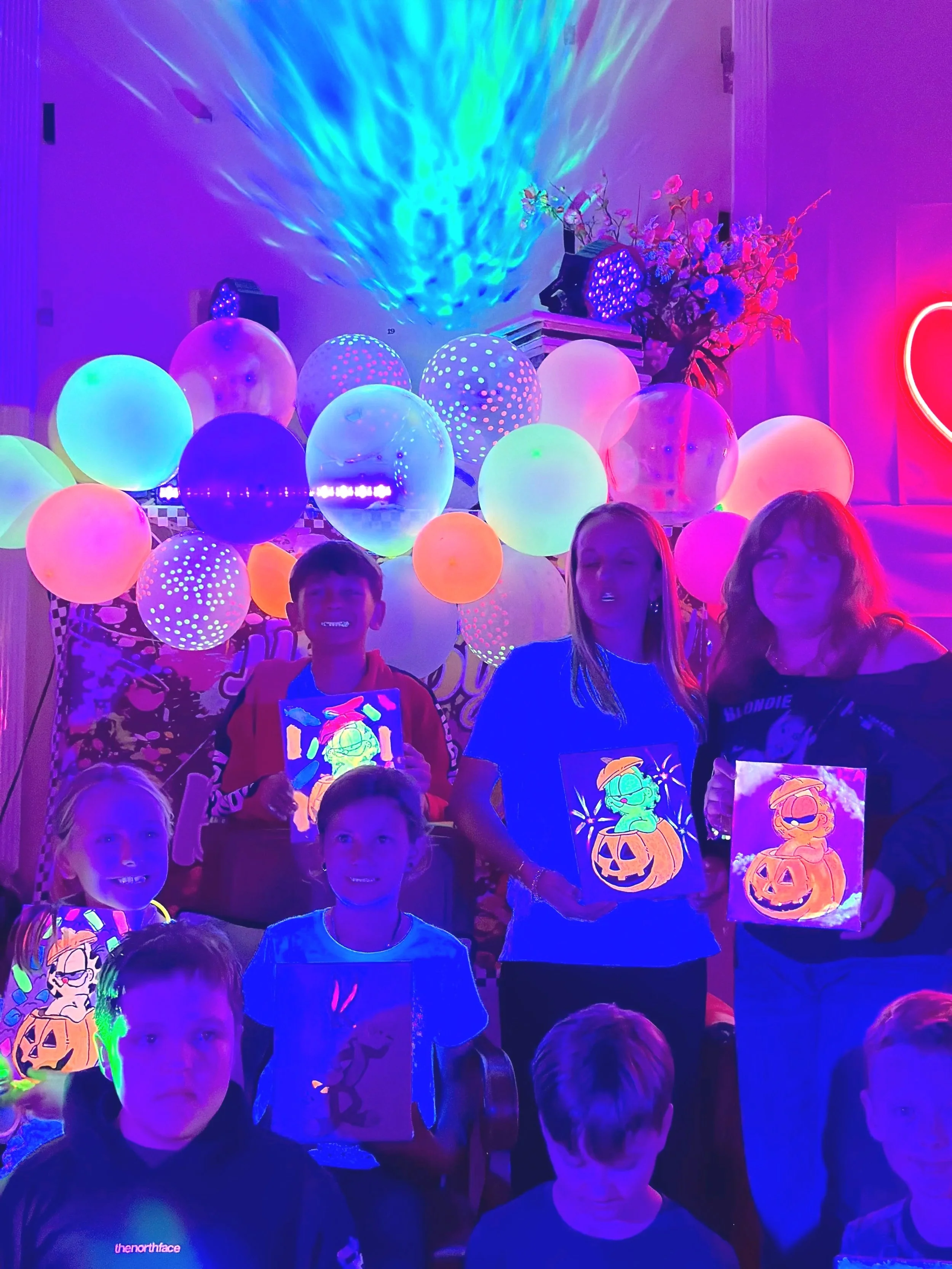 Tweens and teenagers showing their finished glow halloween paintings at the end of a glow art birthday party with light projections, glowing decorations, balloons, and cool lasers.
