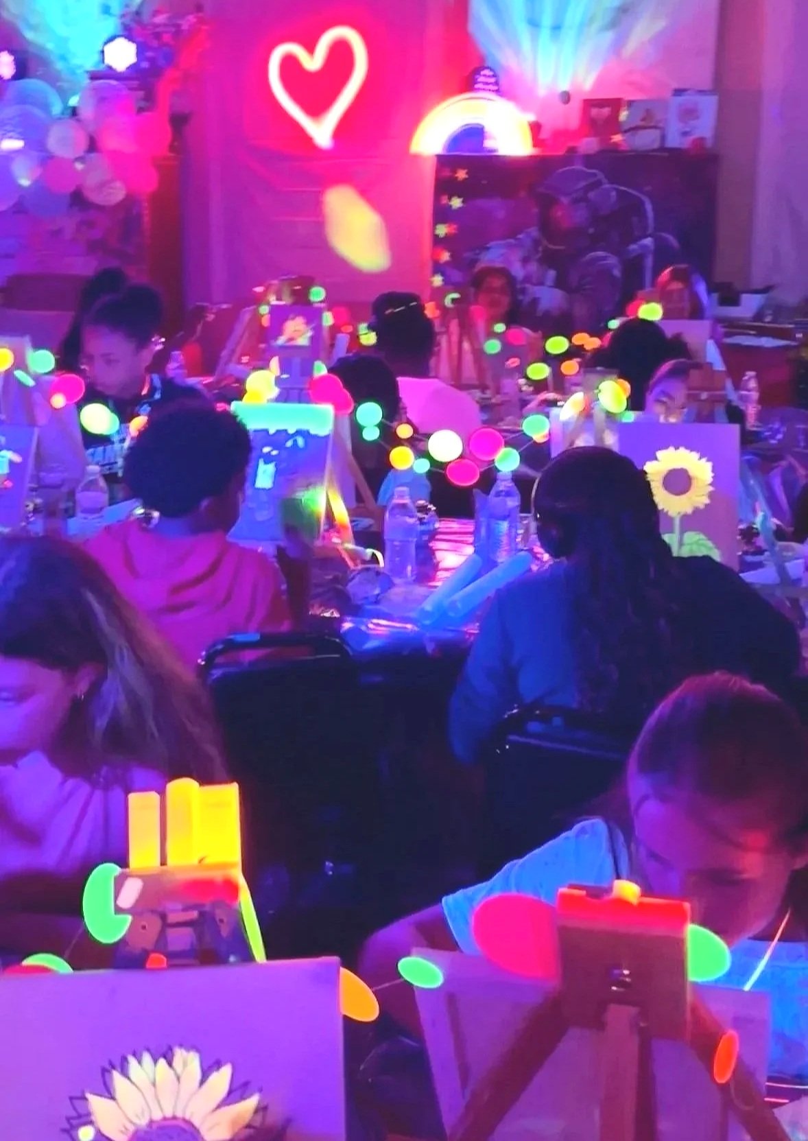 Kids painting at a glow paint birthday party setup with easels, blacklights, balloons, light projections, lights, and neon decorations