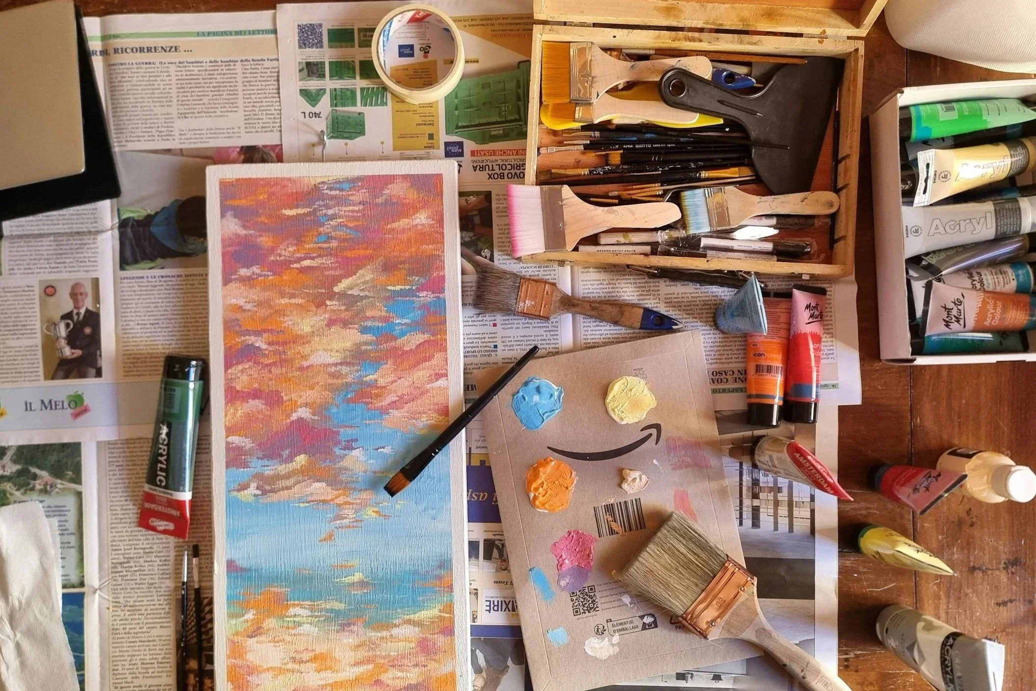 Overhead view of a painting in progress surrounded by brushes, paint, and art supplies on newspaper in a studio setting.