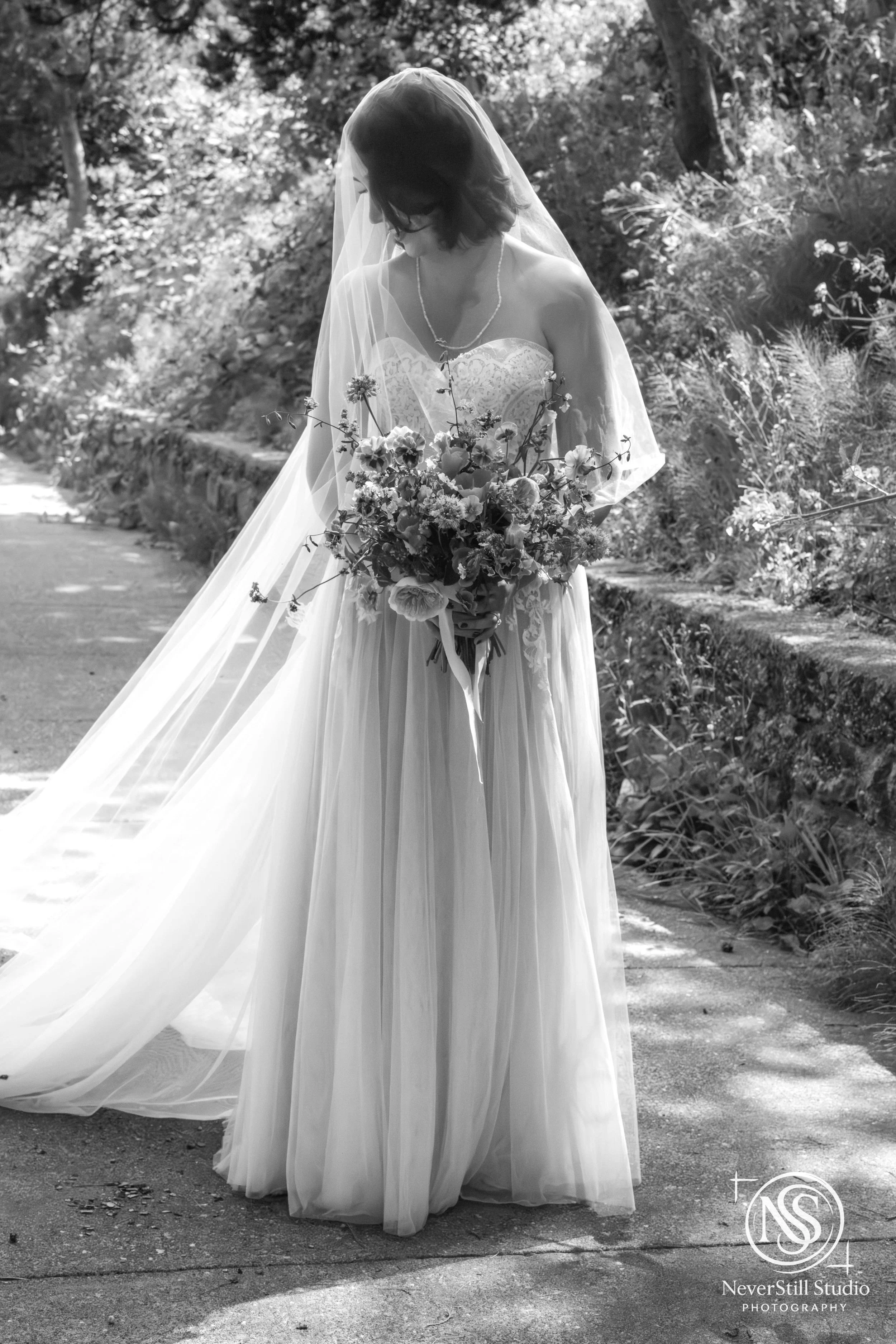 Black and white photo of a bride in a wedding dress holding a bouquet of flowers, standing outdoors on a garden path close to a lake in Washington State.