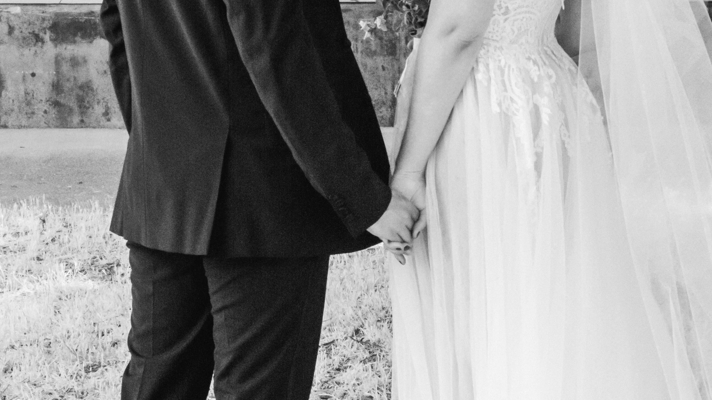 A black and white photograph of a couple holding hands, wearing wedding attire, outdoors on a grassy area near a brick wall outside of a boathouse reception.