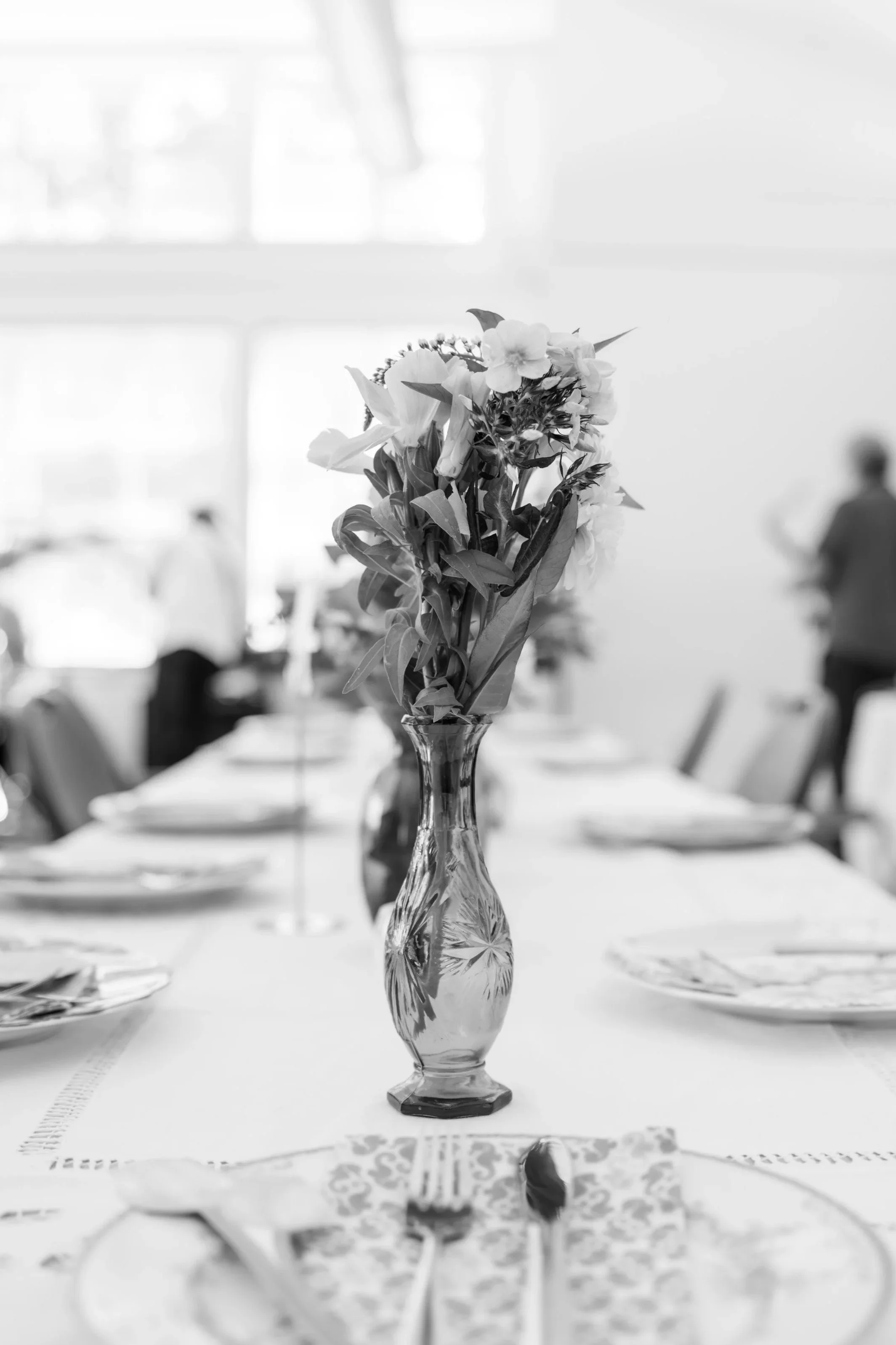 A black and white photo of a dining table with a tall glass vase holding flowers as a centerpiece at a bright and airy wedding.