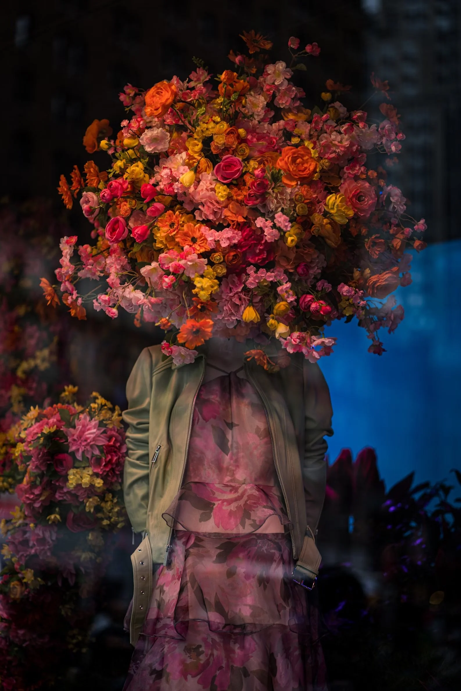 Person with a large bouquet of pink, orange, and yellow flowers obscuring their face, wearing a patterned pink dress and khaki jacket, standing outside at night.