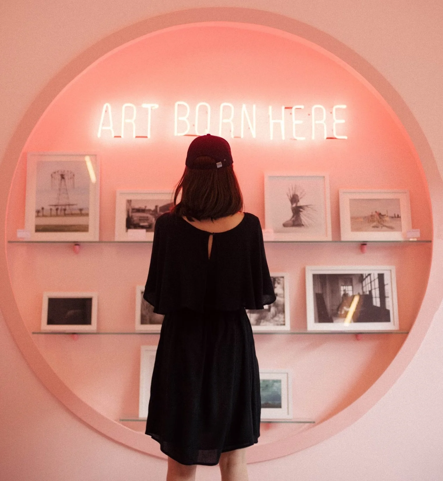 A woman wearing a black dress and cap is looking at framed photographs on a pink wall with a neon sign that says "Art Born Here."