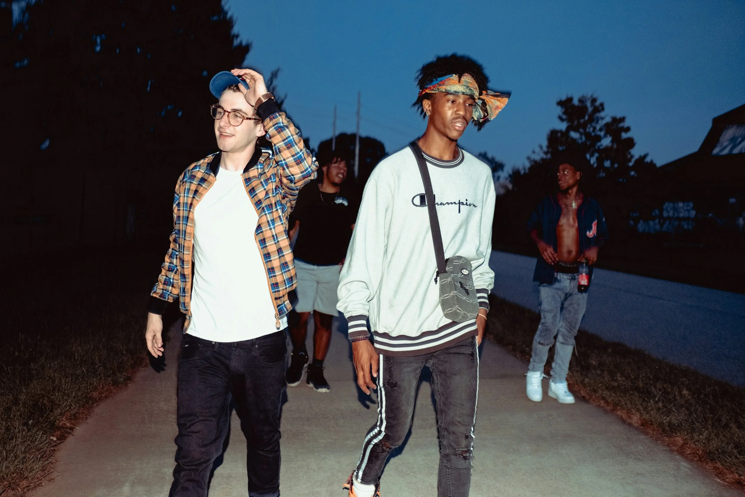 Group of friends walking on a sidewalk at dusk, with one wearing a plaid shirt and cap, and another in a Champion sweatshirt.