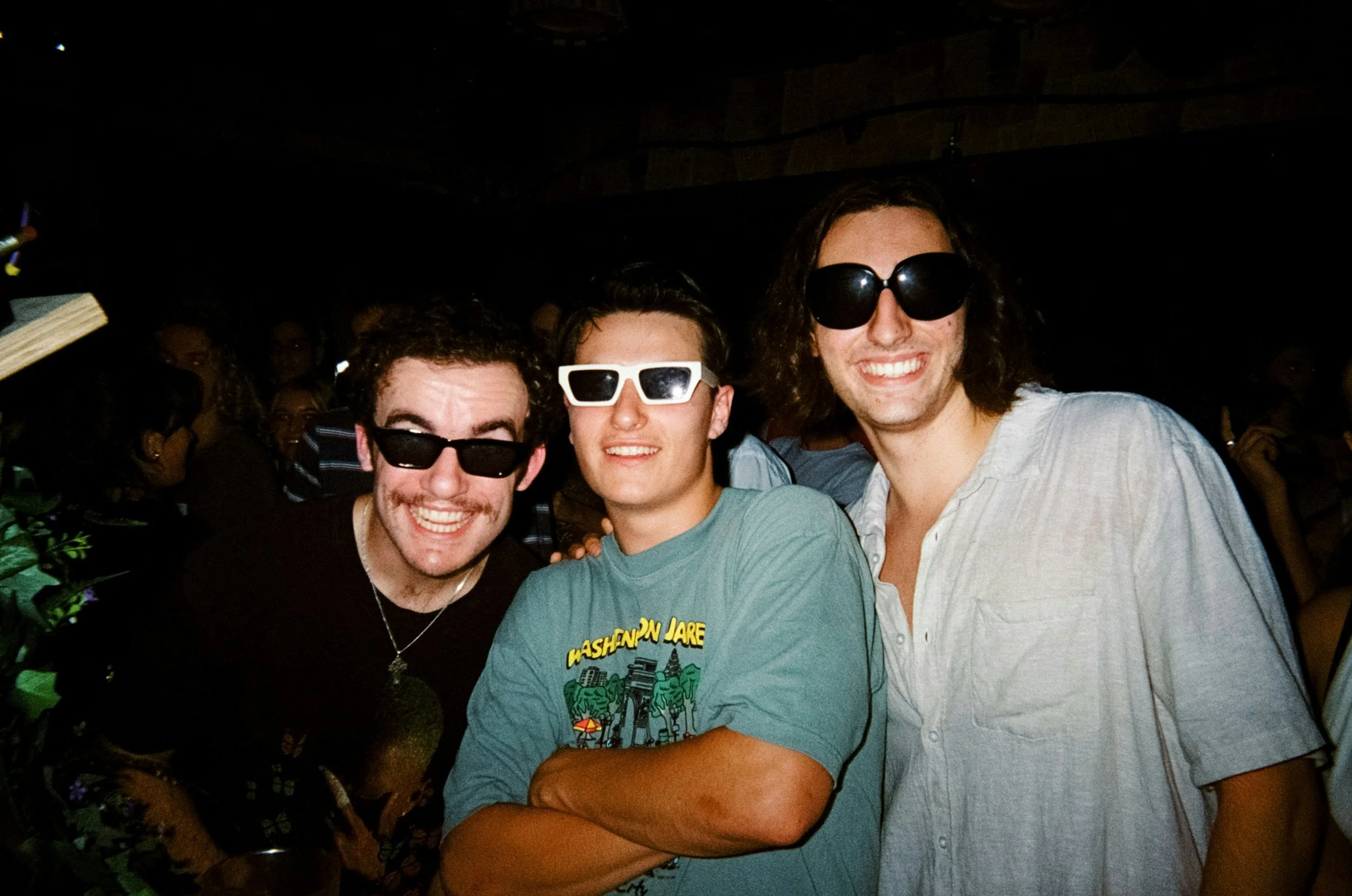 Three people at a party wearing sunglasses, smiling widely. The person on the left has curly hair and a mustache, the middle person has a buzz cut, and the person on the right has long hair. They are surrounded by other partygoers in a dimly lit room.