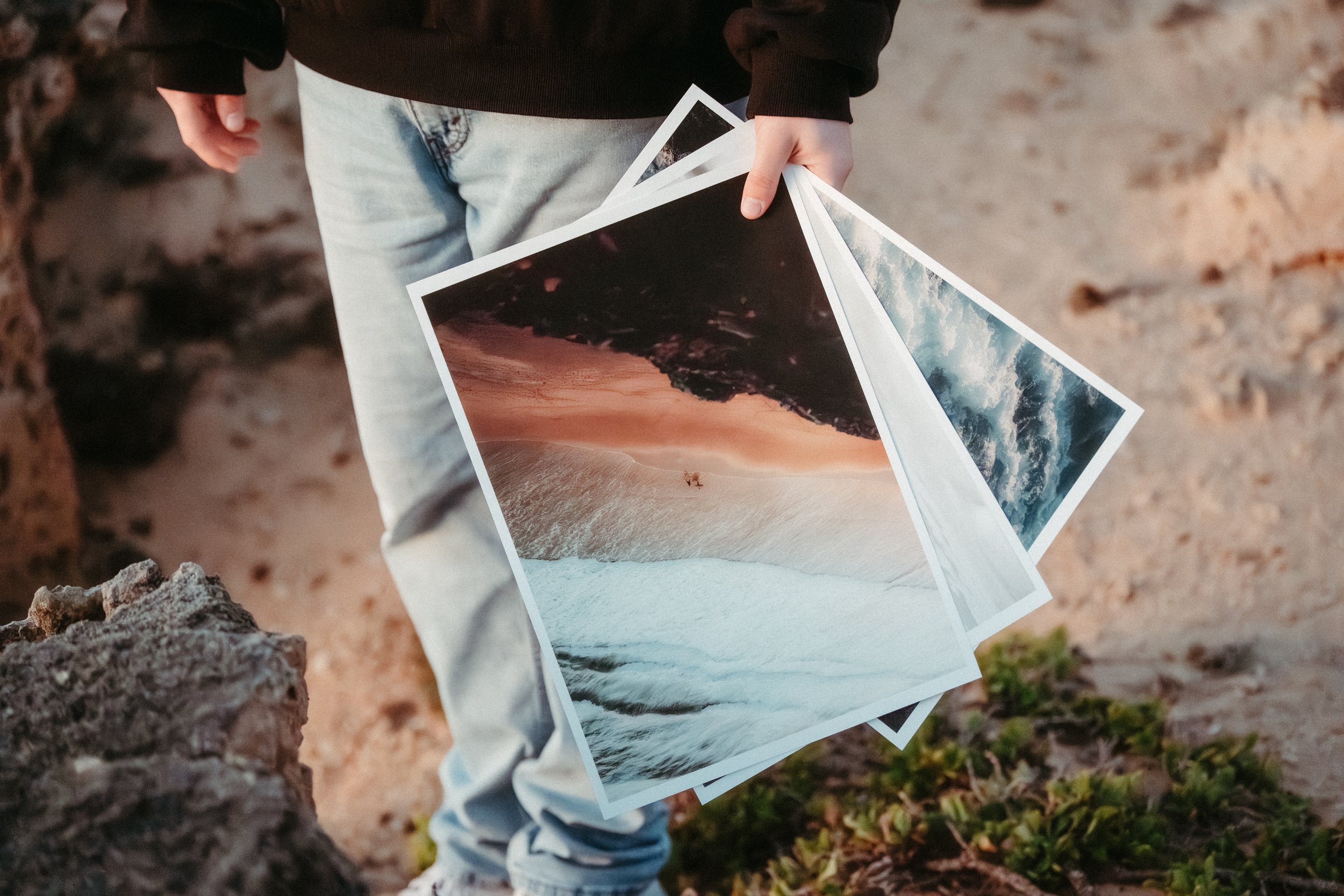 Person holding printed photographs of beach and ocean scenes in a sandy outdoor setting