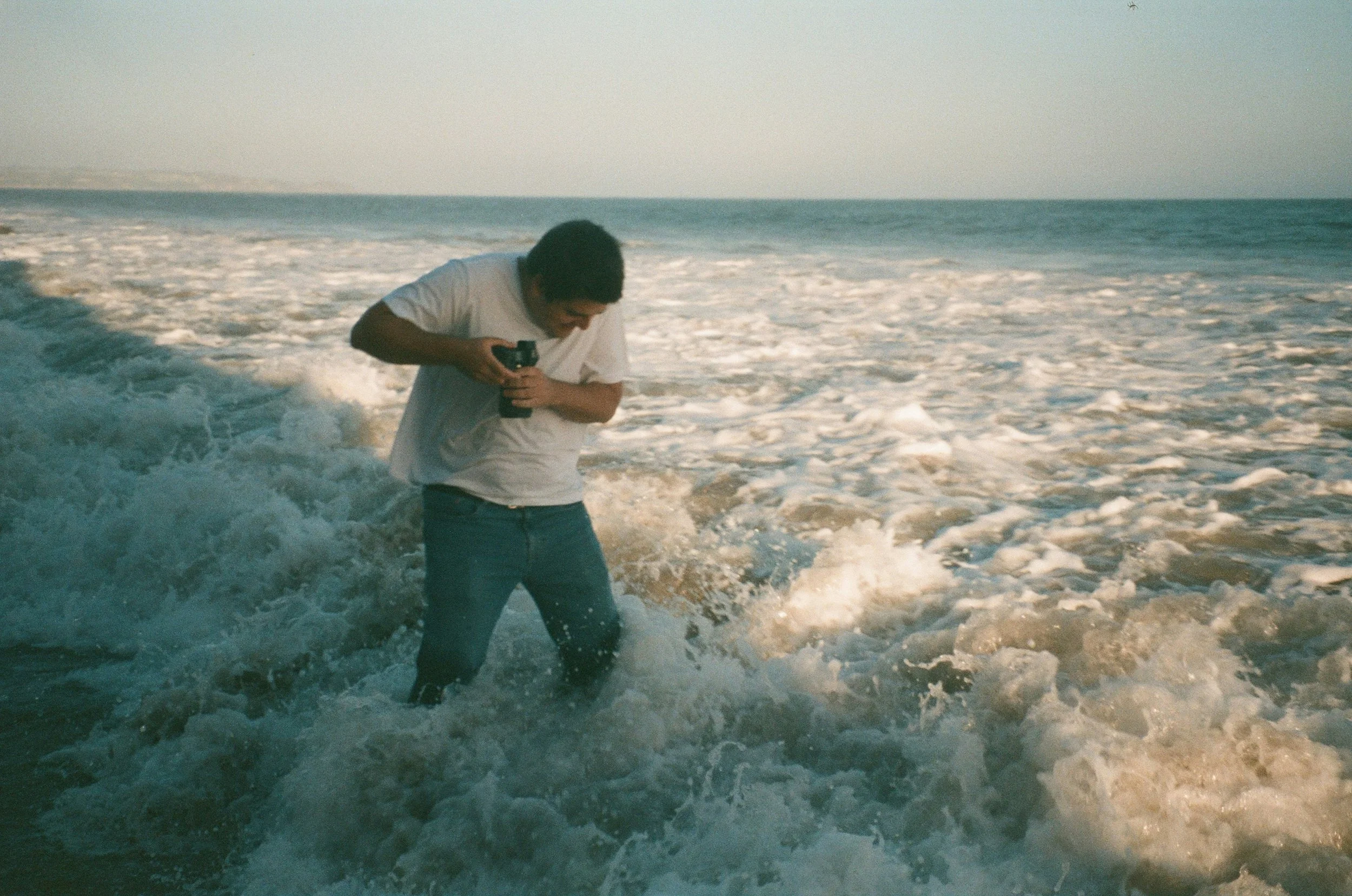 Person standing in ocean waves holding a camera, wearing a white shirt and jeans.