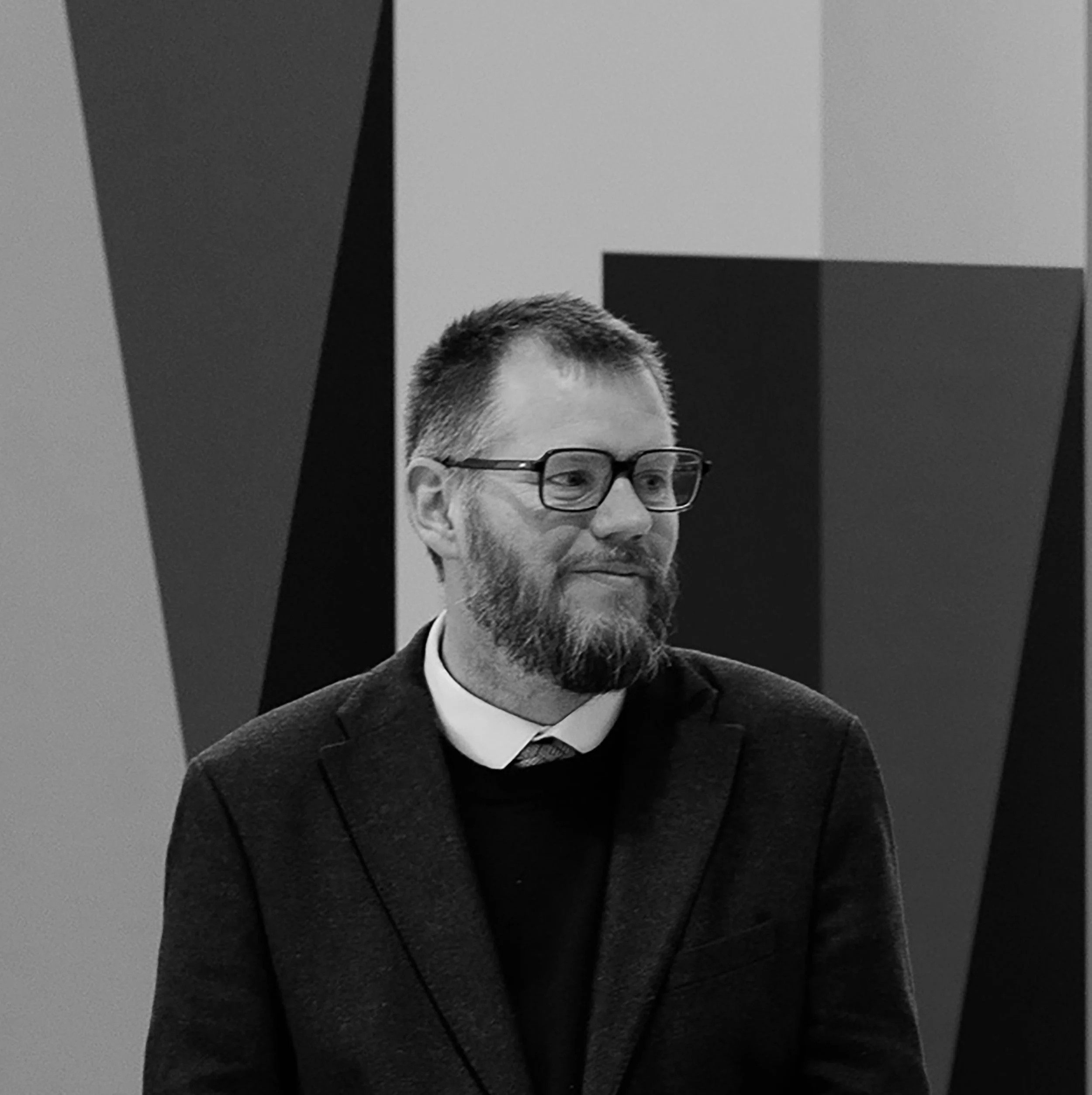 Et svart-hvitt bilde av en mann med briller, skjegg og dress, som står foran abstrakte formasjoner på en vegg.