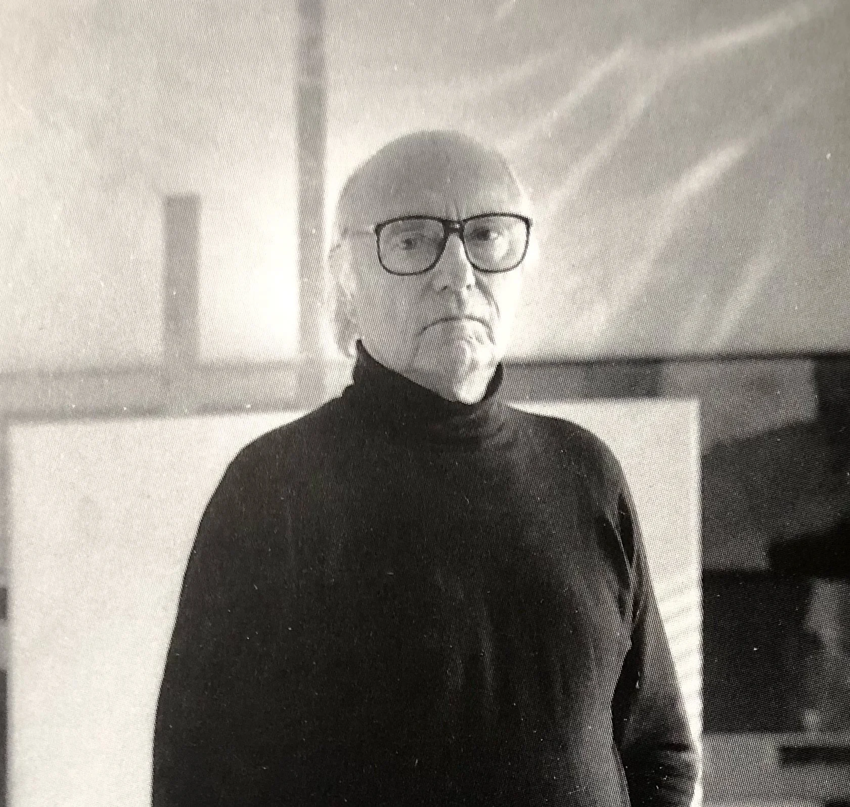 En eldre mann med blankt hår, store svarte briller og svart turtleneck, står foran en vegg med to vaser i bakgrunnen, i svart-hvitt.