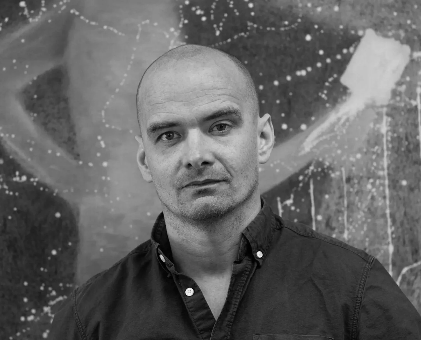 Et svart-hvitt bilde av en mann med skallet hode, iført en knappestolpe skjorte, foran en abstrakt maleri med lyse prikker og striper.