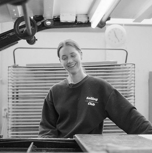 Ung kvinne som smiler, sittende ved et piano i et rom med en bakgrunn av en metallgittervegg.