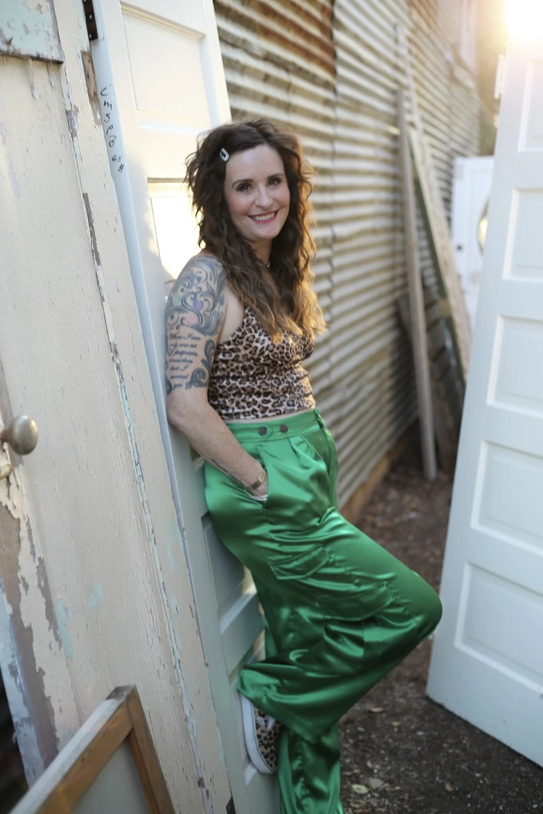 Woman with curly hair wearing a leopard print top, green satin pants, and sneakers, standing outdoors with a corrugated metal wall and doors in the background.