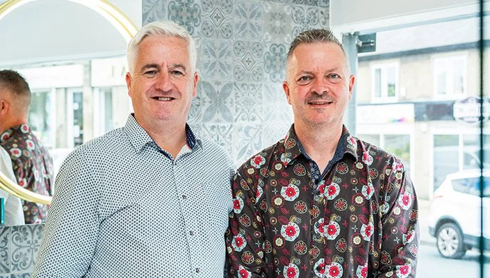 Two middle-aged men standing side by side indoors, smiling at the camera. The man on the left has white hair and wears a white, dotted dress shirt. The man on the right has gray hair and wears a colorful, floral-patterned shirt. A mirror and large window are visible in the background.