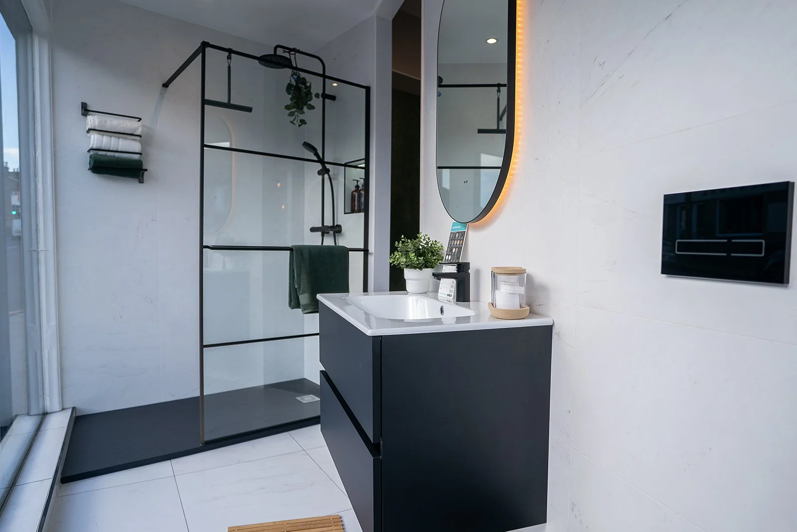 Modern bathroom with a walk-in shower, white and black vanity, round mirror with LED light, and various toiletries.