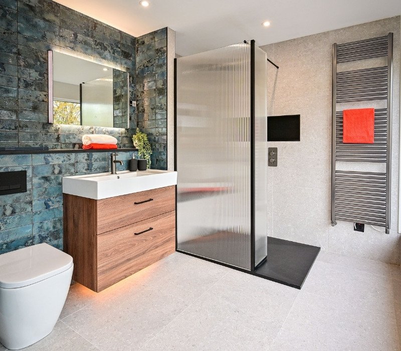 Modern bathroom with a wall-mounted mirror, a rolled towel, a vanity with a sink, a plant, a frosted glass shower enclosure, a wall-mounted TV, and a heated towel rack with a red towel.