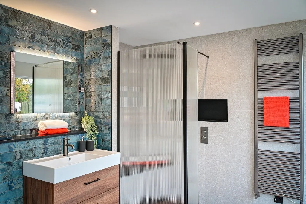 Modern bathroom with textured blue tiles, a large mirror, a white sink with wooden cabinet, and a frosted glass shower enclosure. There are towels and greenery on the shelf, and a black towel warmer with a red towel.