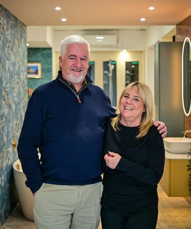 A smiling man with white hair and beard wearing a navy blue sweater, standing next to a smiling woman with blonde hair in a black top; they are indoors in a room with a bathtub, sink, and mirror.