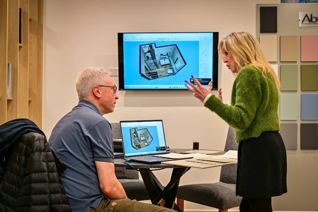 A woman presenting a 3D interior design to a man in an office. The woman is gesturing with her hands, and there is a laptop and monitor showing the same 3D design of a room.