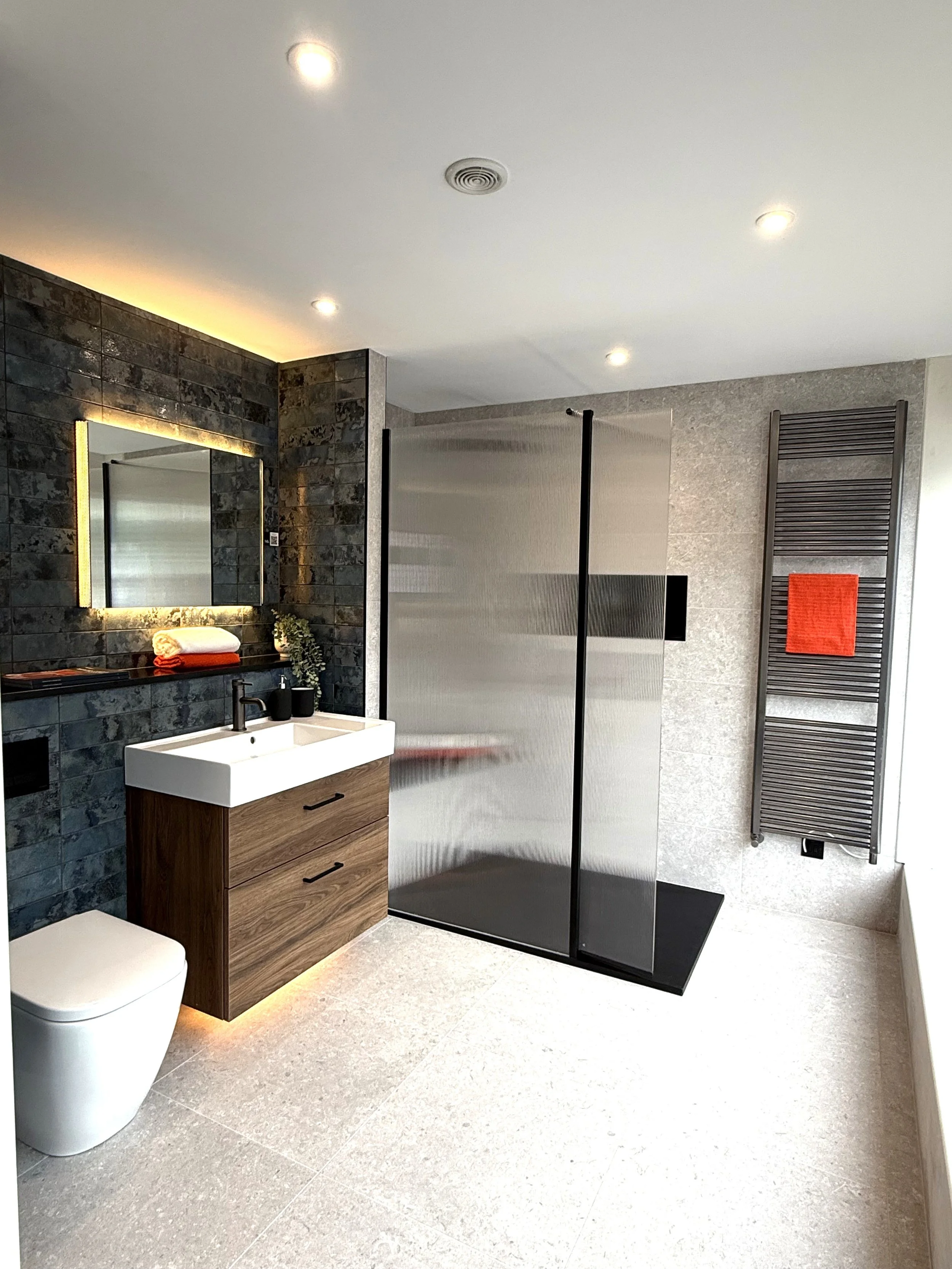 Modern bathroom with a white toilet, a wooden vanity with a white sink, black faucet, black decor, and a large mirror with backlit lighting. There is a textured black tile wall behind the mirror, a frosted glass shower enclosure, and a tall, dark towel warmer with an orange towel. The bathroom features a light-colored tiled floor and ceiling with recessed lighting.