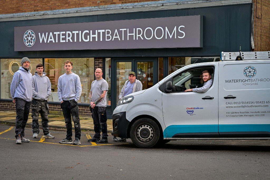 Group of six men standing in front of a business called 'Watertight Bathrooms' with a van parked beside them. The men are wearing casual work clothes, and the van displays company contact information.