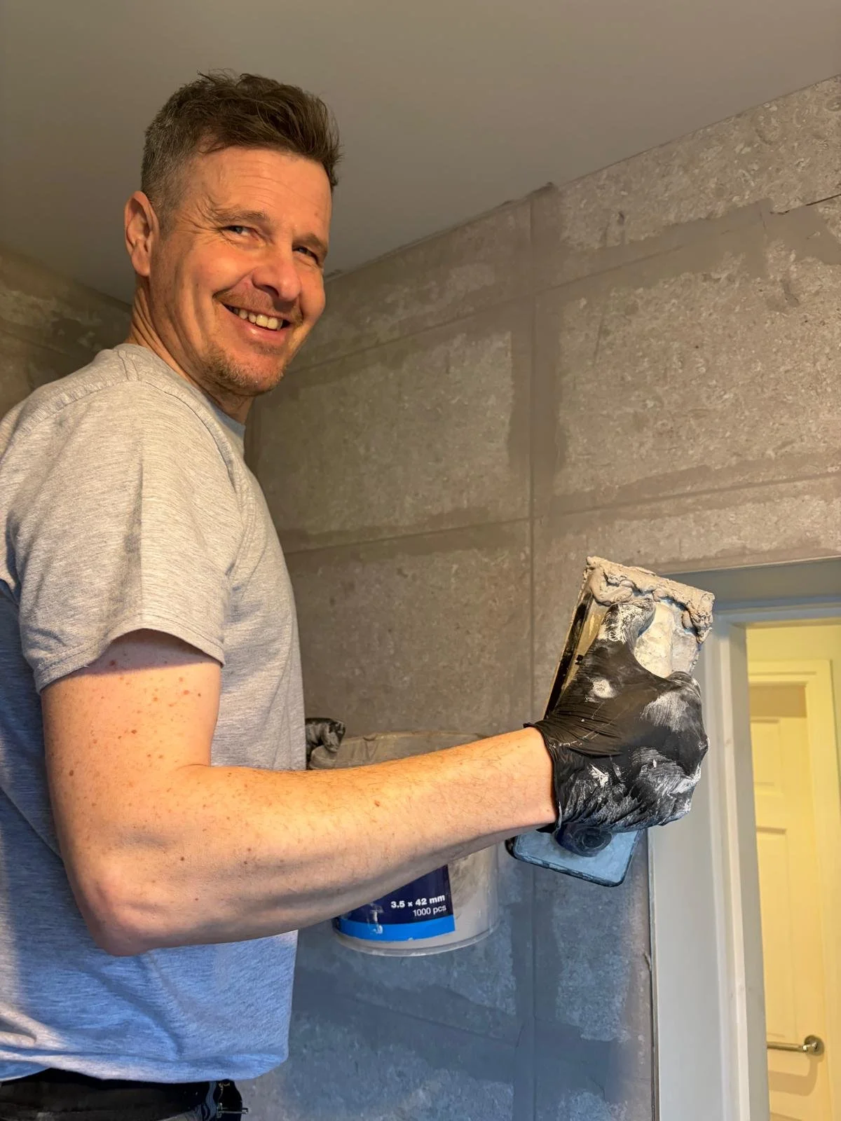 A young man with light skin, short brown hair, and blue eyes, wearing a gray hoodie and a navy blue vest with a logo and text, standing against a plain background.