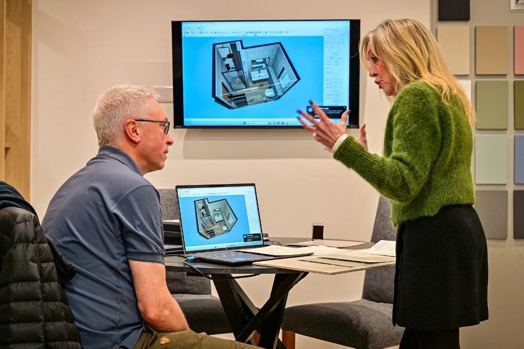 Two people in a meeting room during a design review, with a desktop computer and a large screen displaying 3D interior design plans.