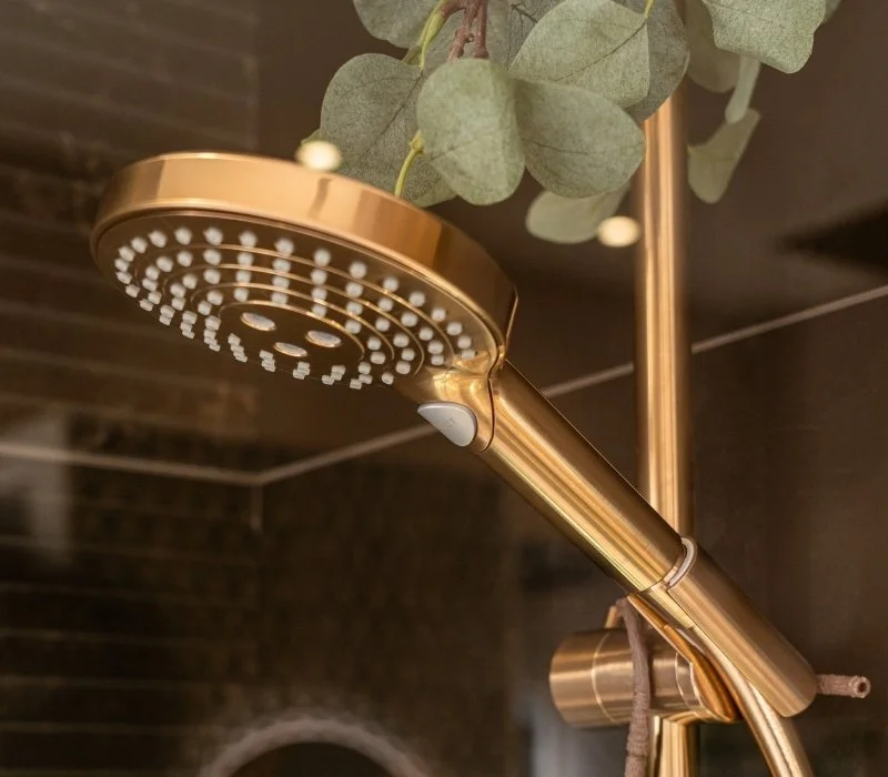 A rose gold showerhead with eucalyptus leaves hanging above in a bathroom with dark tiled walls.