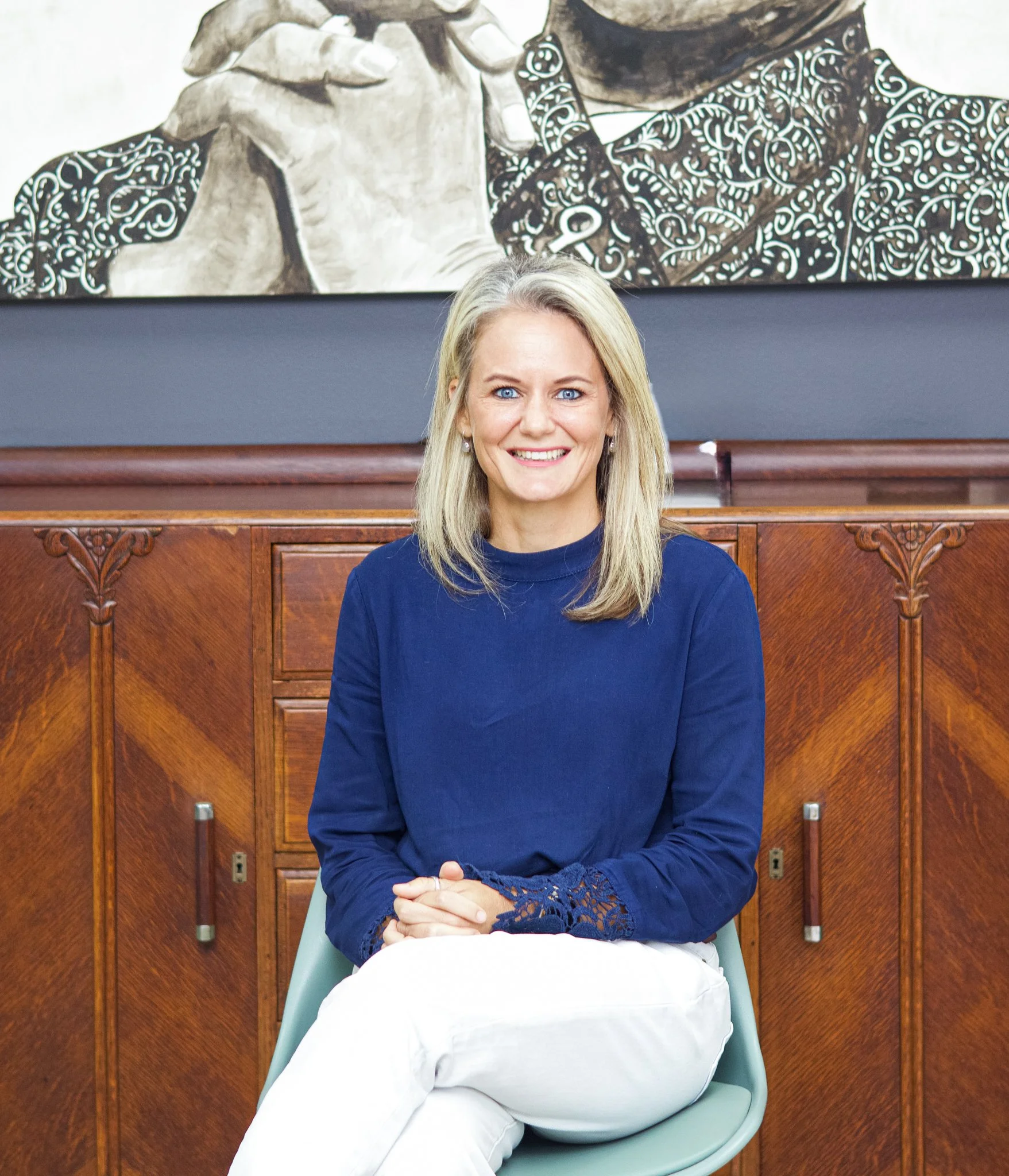 A woman with blonde hair and blue eyes smiling, sitting with her hands clasped in her lap, in front of wooden cabinets and a large painting of a person playing a musical instrument.