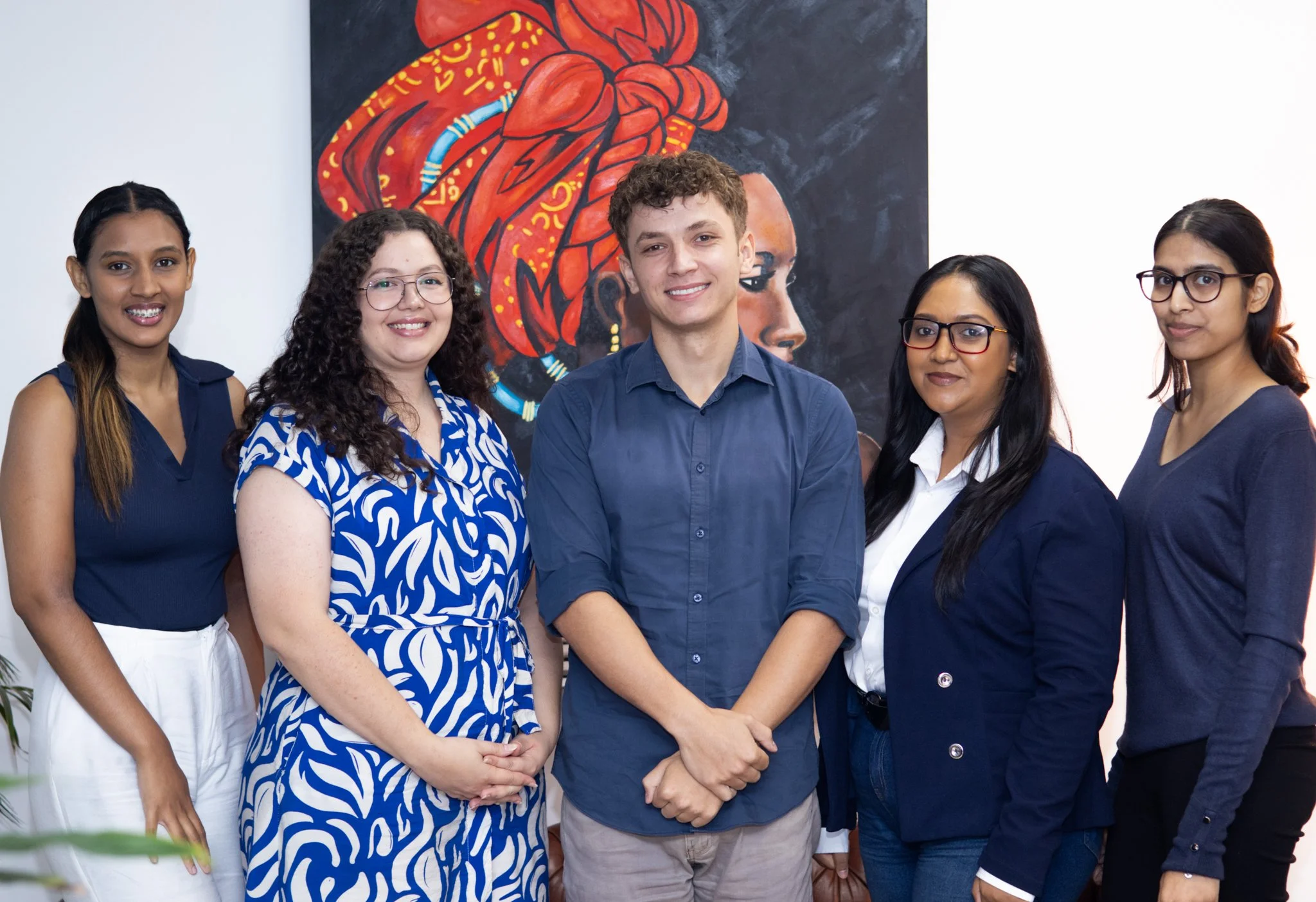 Group of five diverse people standing in front of a colorful painting of a person's profile with a red head wrap, smiling at the camera.